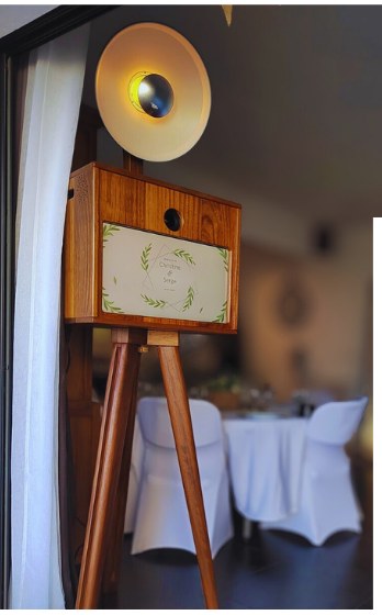 La borne photo en iroko sur son trépied, installée en salle de réception : anneau lumineux allumé, cartel au prénom des mariés, tables dressées derrière